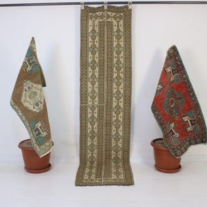 May include: An assortment of vintage rugs and textiles. A long, rectangular rug with a central design is flanked by two smaller rugs draped over terracotta pots. The rugs feature intricate patterns in earthy tones of brown, beige, and red.