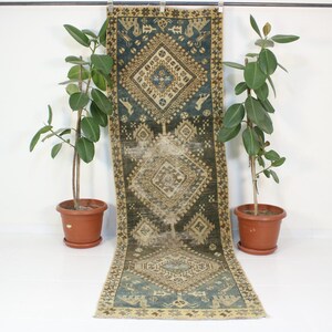 May include: A vintage runner rug with a geometric pattern in shades of blue, brown, and beige. The rug is hanging on a wall with two potted plants on either side.