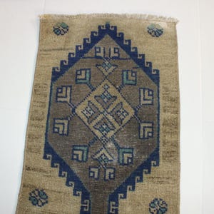 May include: A beige rug with a blue and brown geometric pattern. The rug has a fringed edge.