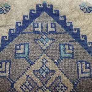 May include: A beige rug with a blue and gray geometric pattern. The pattern features a central diamond shape with smaller diamond shapes surrounding it. The rug has a fringed edge.