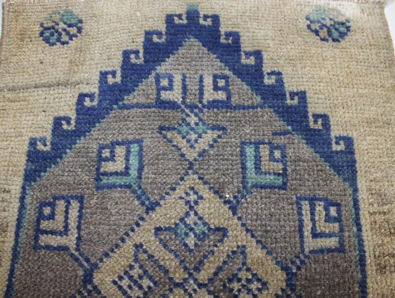 May include: A beige rug with a blue and gray geometric pattern. The pattern features a central diamond shape with smaller diamond shapes surrounding it. The rug has a fringed edge.