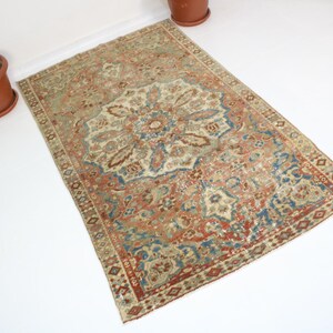 May include: An antique Persian rug with a central medallion design. The rug features a cream-colored field with a detailed floral pattern in shades of red, blue, and brown. The border is a darker red with geometric patterns. The rug is rectangular.