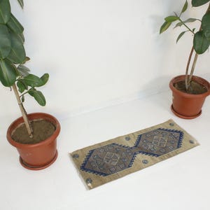 May include: A beige and blue patterned rug with a geometric design. The rug is on a white floor with two potted plants in the background.
