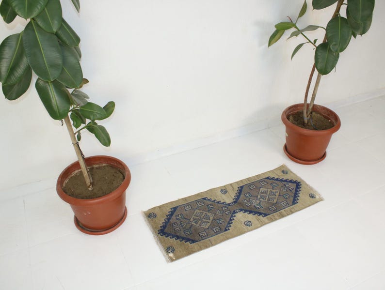 May include: A beige and blue patterned rug with a geometric design. The rug is on a white floor with two potted plants in the background.