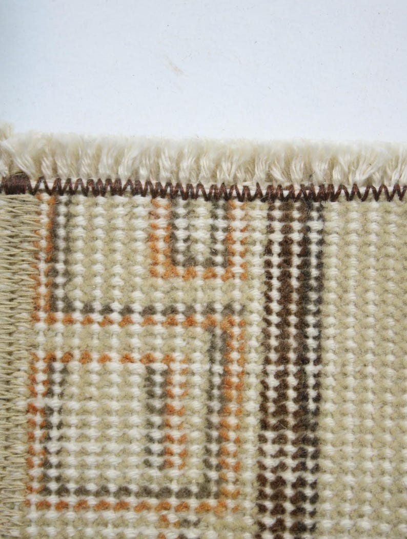 Pode incluir: Close-up de uma se&ccedil;&atilde;o de um tapete tecido com franja creme. O tapete apresenta um padr&atilde;o geom&eacute;trico em tons de marrom e laranja sobre um fundo bege. A textura &eacute; vis&iacute;vel, mostrando a constru&ccedil;&atilde;o tecida.
