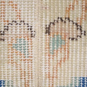 Pode incluir: Close-up de uma se&ccedil;&atilde;o de um tapete vintage. O tapete apresenta um fundo bege com um padr&atilde;o repetitivo de pequenos quadrados. O design inclui detalhes em marrom, laranja, azul e verde-azulado, com uma costura central.