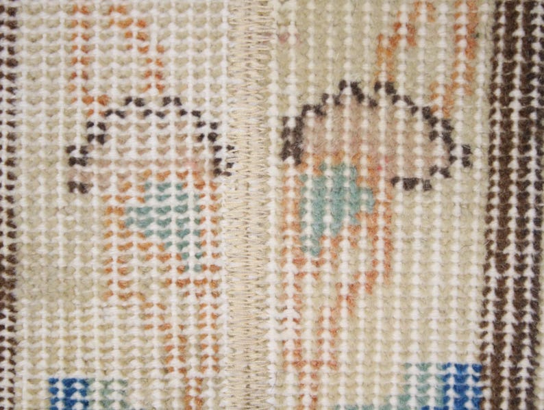 Pode incluir: Close-up de uma se&ccedil;&atilde;o de um tapete vintage. O tapete apresenta um fundo bege com um padr&atilde;o repetitivo de pequenos quadrados. O design inclui detalhes em marrom, laranja, azul e verde-azulado, com uma costura central.