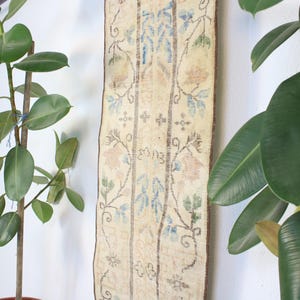 Pode incluir: Um tapete retangular vintage com fundo creme e padr&otilde;es florais em azul, p&ecirc;ssego e verde. O tapete est&aacute; pendurado em uma parede branca, ladeado por plantas em vasos com folhas grandes e verdes.