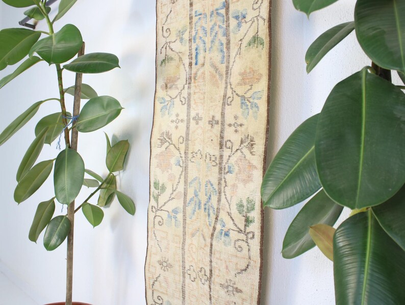 Pode incluir: Um tapete retangular vintage com fundo creme e padr&otilde;es florais em azul, p&ecirc;ssego e verde. O tapete est&aacute; pendurado em uma parede branca, ladeado por plantas em vasos com folhas grandes e verdes.