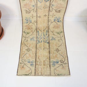 Pode incluir: Um tapete retangular de estilo vintage com fundo bege e padr&otilde;es florais em azul, p&ecirc;ssego e verde. O tapete tem um design central com listras verticais e uma borda marrom escuro. Dois vasos de terracota est&atilde;o em ambos os lados.