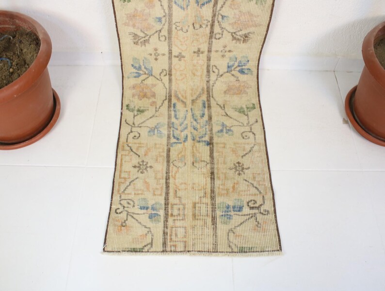 Pode incluir: Um tapete retangular de estilo vintage com fundo bege e padr&otilde;es florais em azul, p&ecirc;ssego e verde. O tapete tem um design central com listras verticais e uma borda marrom escuro. Dois vasos de terracota est&atilde;o em ambos os lados.