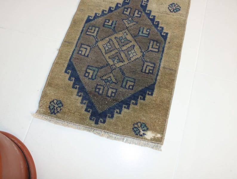 May include: A beige rug with a blue and brown geometric pattern. The rug has a fringe border.