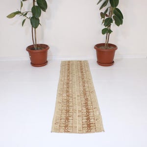 Può includere: Un tappeto da corridoio beige con un motivo geometrico marrone. Il tappeto è steso su un pavimento di piastrelle bianche.