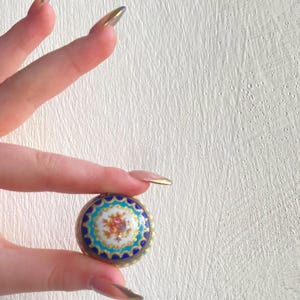 Puede incluir: Una caja redonda de esmalte azul y dorado con un diseño floral. La caja está sostenida en una mano con uñas largas y manicuradas.