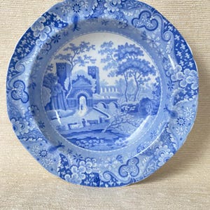 May include: A round, blue and white ceramic plate with a detailed landscape scene in the center. The plate features a floral and scrollwork border. The landscape depicts a bridge, buildings, and trees. The plate is likely vintage.