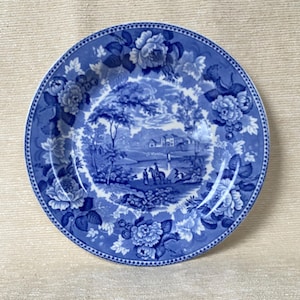 May include: A round blue and white decorative plate with a floral border and a central landscape scene. The plate features a detailed design of flowers and a pastoral landscape with trees, buildings, and figures.