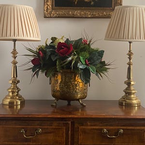 May include: Two brass table lamps with pleated cream shades either side of a decorative brass planter filled with red roses and greenery. The lamps sit on a dark wood dresser with brass hardware, beneath a framed painting.