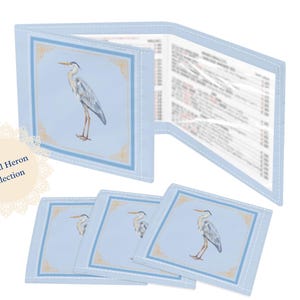 May include: A light blue Coastal Heron Collection set featuring a wallet and coasters. The wallet is open, revealing printed pages. Each item has a heron illustration within a decorative border. The set is on a light blue background.