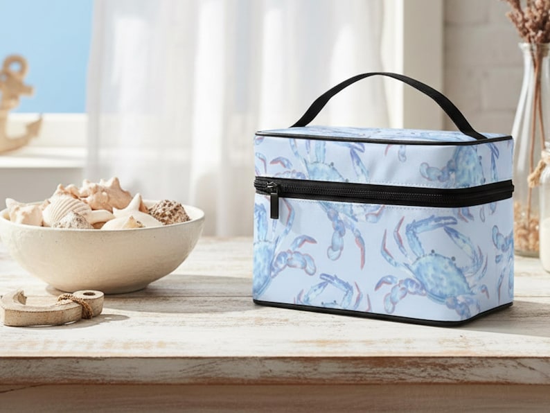 May include: A light blue cosmetic bag with a black zipper and handle. The bag features a repeating pattern of blue watercolor crabs. A bowl of seashells sits on a wooden table.