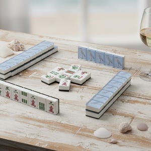 May include: A Mahjong set with white tiles, blue accents, and Chinese characters is arranged on a weathered wooden table. A glass of white wine and seashells are also present. The set includes racks and individual tiles.