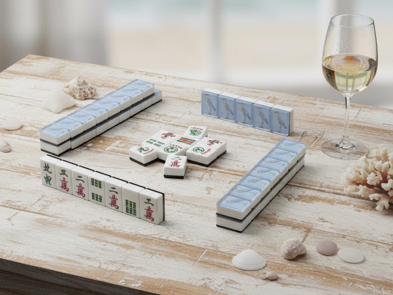 May include: A Mahjong set with white tiles, blue accents, and Chinese characters is arranged on a weathered wooden table. A glass of white wine and seashells are also present. The set includes racks and individual tiles.