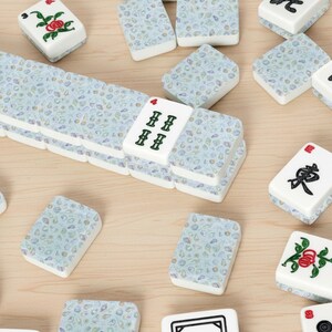 May include: A collection of white and blue Mahjong tiles scattered on a light wood surface. The tiles feature various symbols, numbers, and floral designs in red, green, and black. Some tiles are stacked, while others are scattered.