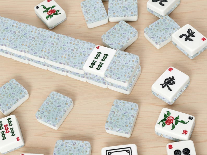 May include: A collection of white and blue Mahjong tiles scattered on a light wood surface. The tiles feature various symbols, numbers, and floral designs in red, green, and black. Some tiles are stacked, while others are scattered.