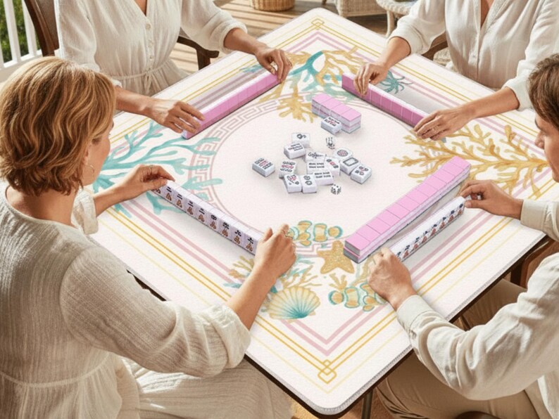 May include: A square Mahjong table with a cream-colored surface featuring a coral reef design in teal, gold, and pink. Four people are playing Mahjong, with pink tile racks and white tiles visible. The game is set on a wooden table.