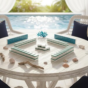 May include: A round, white wooden table set for a game of Mahjong. The table is surrounded by two white wicker chairs with navy blue cushions. Champagne glasses and decorative seashells are also on the table. The game pieces are white with teal and green accents.