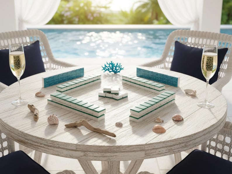 May include: A round, white wooden table set for a game of Mahjong. The table is surrounded by two white wicker chairs with navy blue cushions. Champagne glasses and decorative seashells are also on the table. The game pieces are white with teal and green accents.