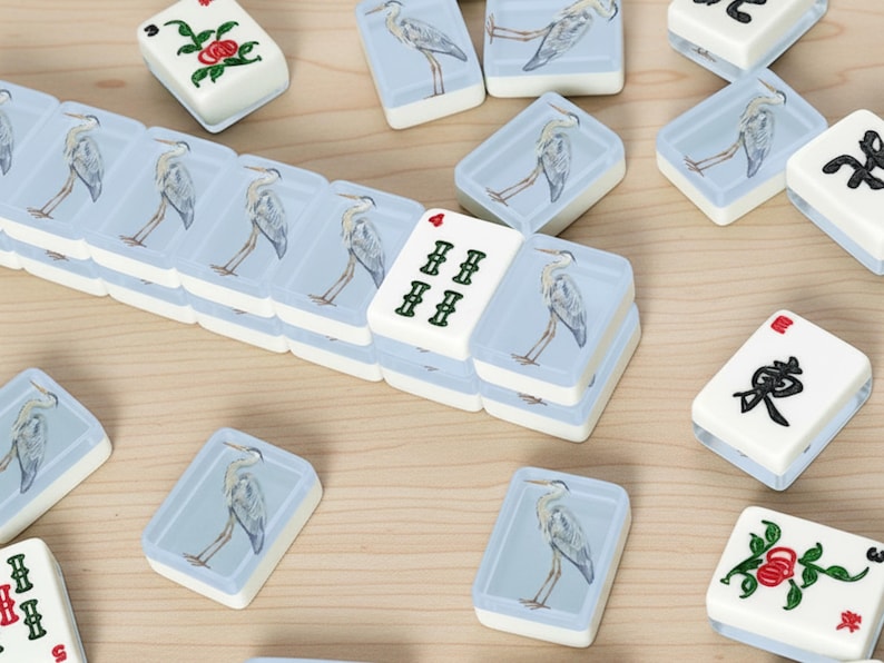 May include: A collection of white and blue Mahjong tiles. The tiles feature illustrations of a heron and floral designs. Some tiles display Chinese characters and numbers. The tiles are arranged on a light brown surface.
