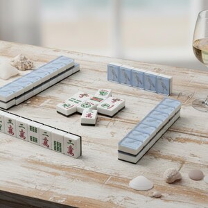 May include: A Mahjong set with white tiles, blue accents, and Chinese characters is arranged on a weathered wooden table. A glass of white wine and seashells are also present. The set includes racks and individual tiles.