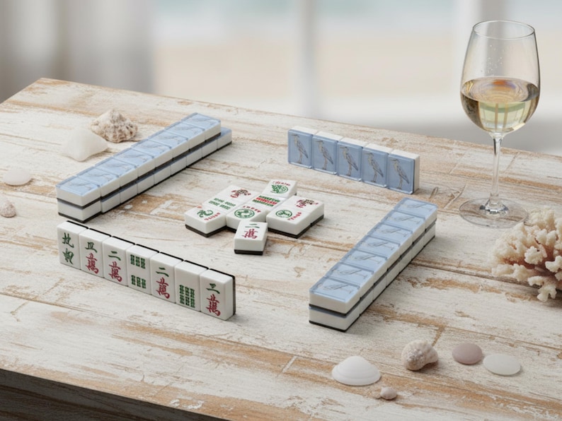 May include: A Mahjong set with white tiles, blue accents, and Chinese characters is arranged on a weathered wooden table. A glass of white wine and seashells are also present. The set includes racks and individual tiles.
