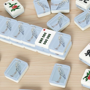 May include: A collection of white and blue Mahjong tiles. The tiles feature illustrations of a heron and floral designs. Some tiles display Chinese characters and numbers. The tiles are arranged on a light brown surface.