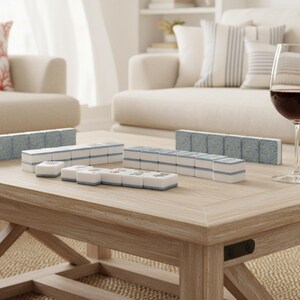 May include: A light wood coffee table displays a Mahjong set with blue and white tiles. A glass of red wine sits nearby. The game set is arranged on the table, ready for play. The room has a neutral color palette.