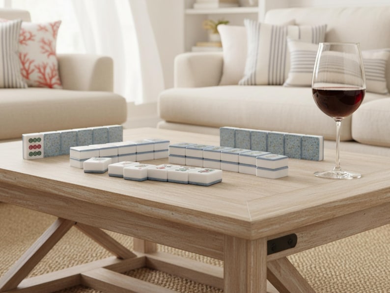 May include: A light wood coffee table displays a Mahjong set with blue and white tiles. A glass of red wine sits nearby. The game set is arranged on the table, ready for play. The room has a neutral color palette.