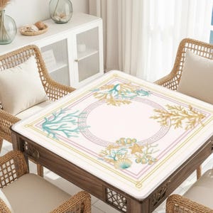 May include: A square wooden table with a decorative, light-colored top featuring a sea-themed design. The design includes coral, seashells, and starfish in shades of blue, gold, and pink. The table is surrounded by four wicker chairs with cream-colored cushions.