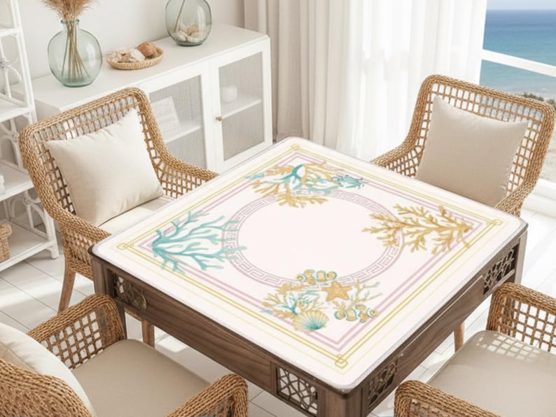 May include: A square wooden table with a decorative, light-colored top featuring a sea-themed design. The design includes coral, seashells, and starfish in shades of blue, gold, and pink. The table is surrounded by four wicker chairs with cream-colored cushions.