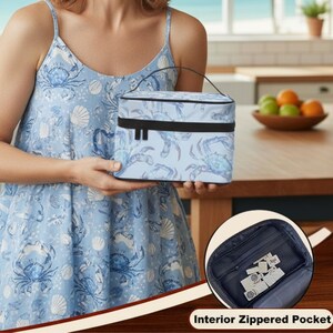 May include: A light blue insulated lunch bag with a crab and seashell pattern. The bag has a black handle and trim. The image also shows a close-up of the interior zippered pocket. The bag is held by a person wearing a blue dress with a matching crab and seashell pattern.