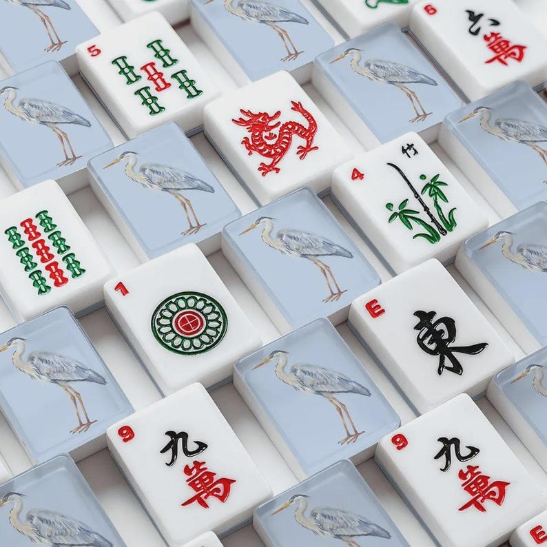 May include: A close-up of a Mahjong set with white tiles. Some tiles feature a heron on a light blue background. Other tiles have red and green symbols, a red dragon, and bamboo. The set is arranged in a grid.