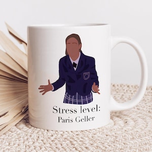 Op de afbeelding: Een witte keramische mok met een cartoonillustratie van een vrouw met bruin haar die een blauwe blazer en een geruite rok draagt. De tekst "Stress level: Paris Geller" staat op de mok gedrukt.
