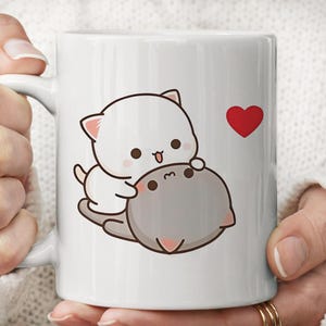 Könnte beinhalten: Ein weißer Keramikbecher mit einer grafischen Illustration einer kleinen weißen Katze, die auf einer größeren grauen Katze sitzt. Die weiße Katze hat rosa Ohren und einen fröhlichen Ausdruck, rechts daneben befindet sich ein kleines rotes Herz.