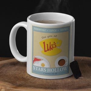 Peut inclure: Une tasse en céramique blanche avec un design de diner rétro comportant le texte "See you at Luke's" et "Stars Hollow, Connecticut". La tasse est de couleur bleu, jaune et rouge et représente une assiette d'œufs et une tasse de café.