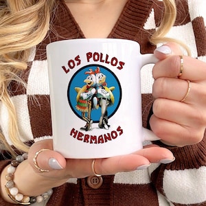 Peut inclure: Une tasse en céramique blanche avec un graphique bleu, rouge et blanc de deux poulets de dessin animé avec le texte "Los Pollos Hermanos".