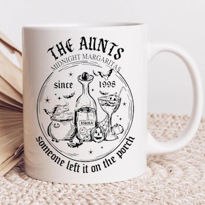 May include: White ceramic mug with a black graphic design. The design features the text "The Aunts Midnight Margaritas since 1998" with illustrations of a cauldron, tequila bottle, cocktail, pumpkins, and bats. The phrase "someone left it on the porch" is also included.