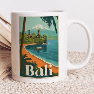 Könnte beinhalten: Weiße Keramiktasse mit einer bunten Illustration eines Tempels in Bali, Indonesien. Das Bild zeigt einen Strand, das Meer, Palmen und einen Berg im Hintergrund. Der Text "Bali" ist in großen weißen Buchstaben auf die Tasse gedruckt.