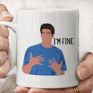 Op de afbeelding: Een witte keramische mok met een grafische illustratie van een persoon met donker haar in een blauw T-shirt, die met beide handen gebaart. De tekst "I'M FINE." staat in zwarte hoofdletters rechts naast de illustratie gedrukt.