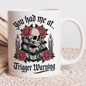 Può includere: Tazza in ceramica bianca con grafica nera e rossa. Il design presenta un teschio, rose, candele e il testo "You had me at... Trigger Warning" in un carattere gotico. La tazza ha un manico curvo.