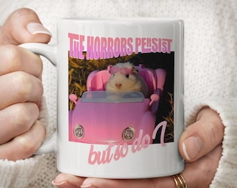 Pode incluir: Uma caneca de cerâmica branca com um gráfico de um porquinho-da-índia branco usando óculos de sol cor-de-rosa enquanto dirige um carro de brinquedo rosa. O texto acima diz THE HORRORS PERSIST e o texto abaixo diz but so do I em rosa.