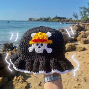 May include: Black crochet bucket hat with a white trim and a yellow and red pirate hat emblem with a skull and crossbones. The hat is handmade and suitable for cosplay or everyday wear.